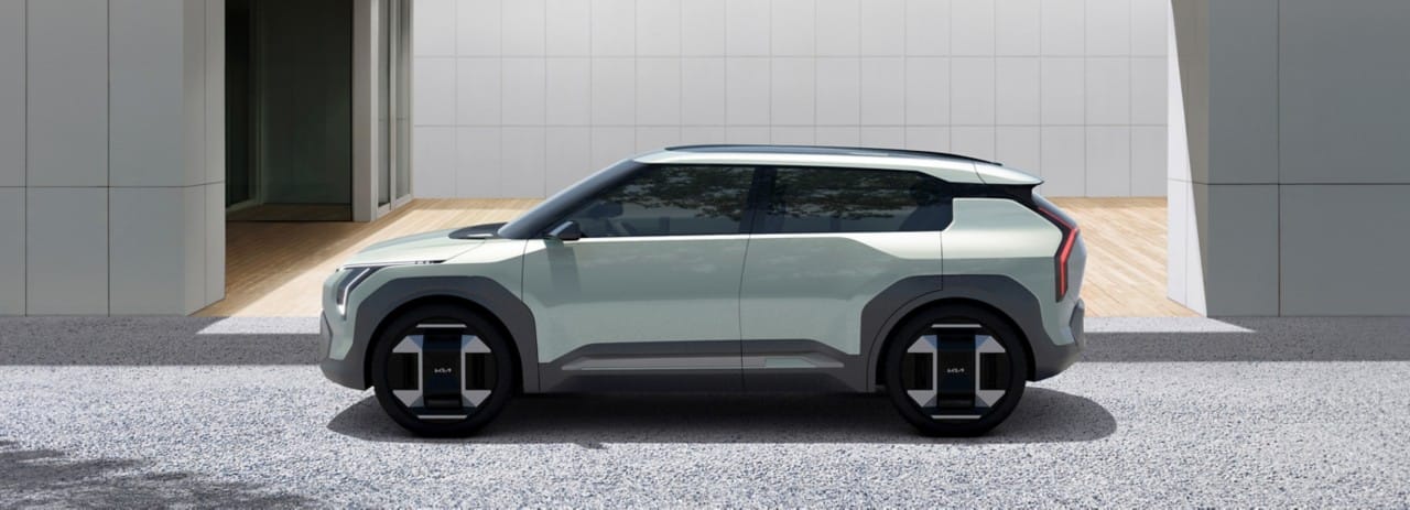 Kia EV3 concept vehicle in light green, side view, parked outside of a building featuring a wrap-around windscreen giving the illusion of a floating roof.