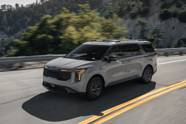 2025 Kia Carnival in gray, driving on an open road on a hill-side highway, front three-quarter view