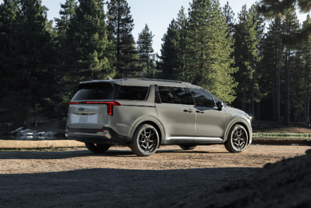 2025 Kia Carnival in gray, parked off-road near a lake, rear three-quarter view